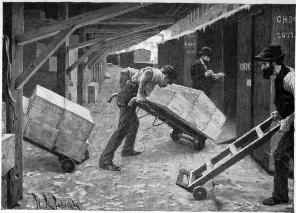 Three men are under cover on a loading dock. There are box cars next to the loading dock. One of the men is pushing a hand truck loaded with crates into one of the boxcars.