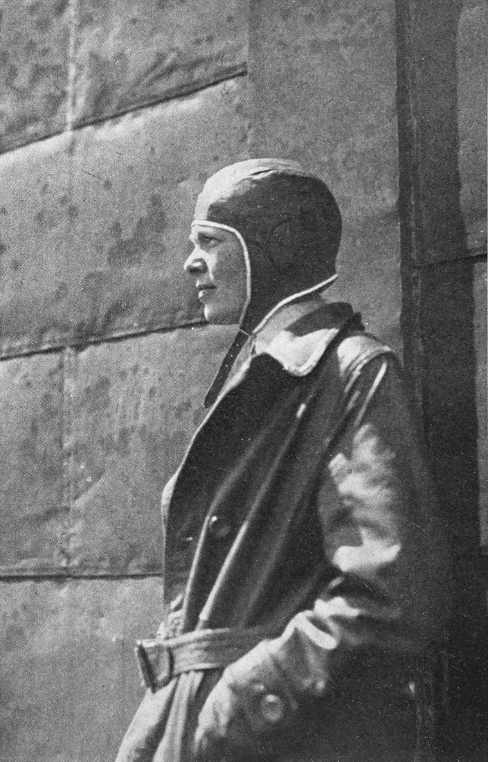 Photo medium profile shot of Earhart wearing flight gear outside a block building