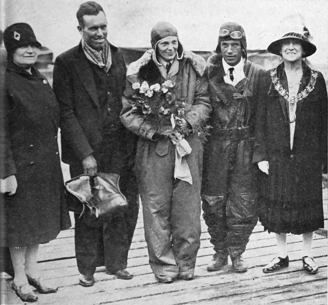Photo wide shot of 5 people; Earhart is in middle holding a bouquet of roses. Earhart and Stultz are wearing flight gear