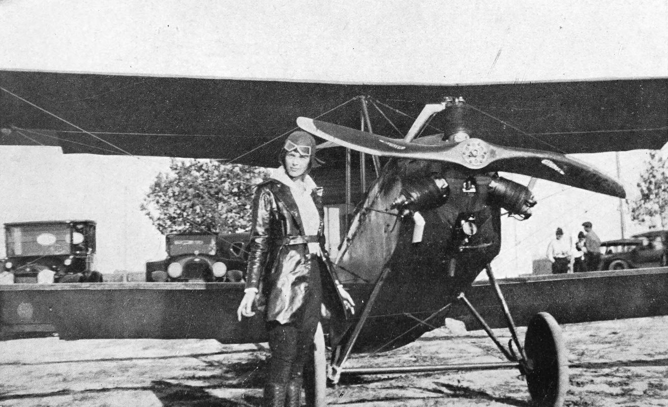 Photo wide shot of Earhart in flight gear standing in front of a biplane