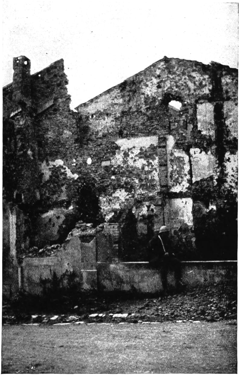 A uniformed man sitting on the remains of a wall with damanged buildings behind.