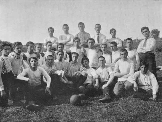 The boys appear to be mostly teenagers. They are arranged in three rows directly facing the camera, with the nearest row seated and the back row standing. There is a ball in front of the boys, and the boys are wearing light tee shirts and dark pants. Two in the back appear to be older and are wearing shirts with ties. There are about twenty-five total individuals.