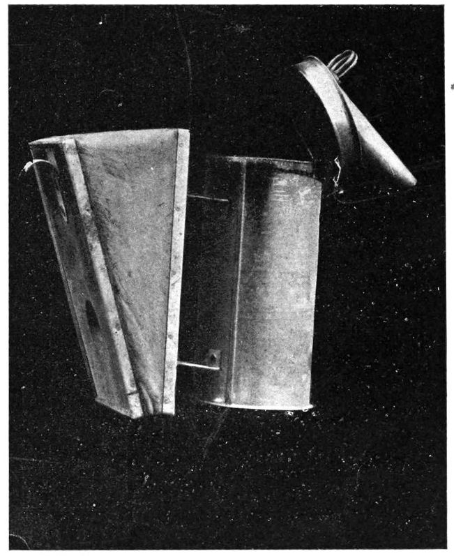 Smoker has a billows on the left, connected to a metal cylinder. A lid for the cylinder with a spout is titled to be open.