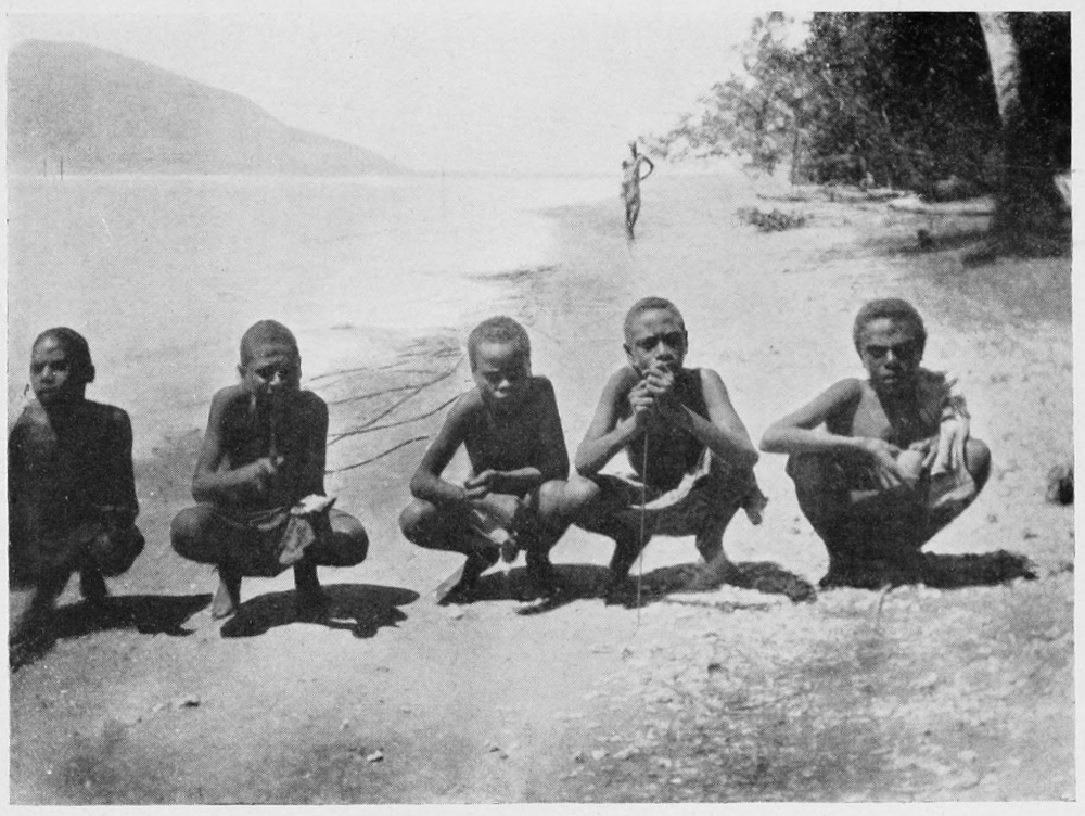 Fivenative men in local garb squat in a row along the shoreline, facing the viewer.