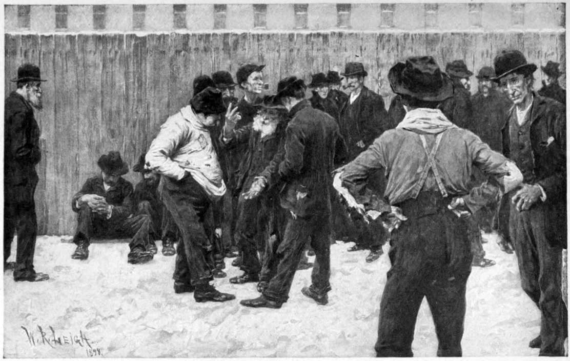 Men gathered outside. A fence
is visible behind them with a wide building visible behind the fence.