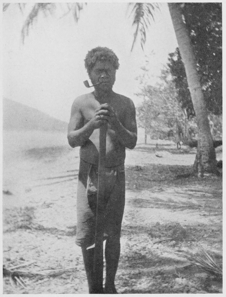 Standing native male, pipe in his mouth, holds a staff in front of him, while standing on the shoreline.